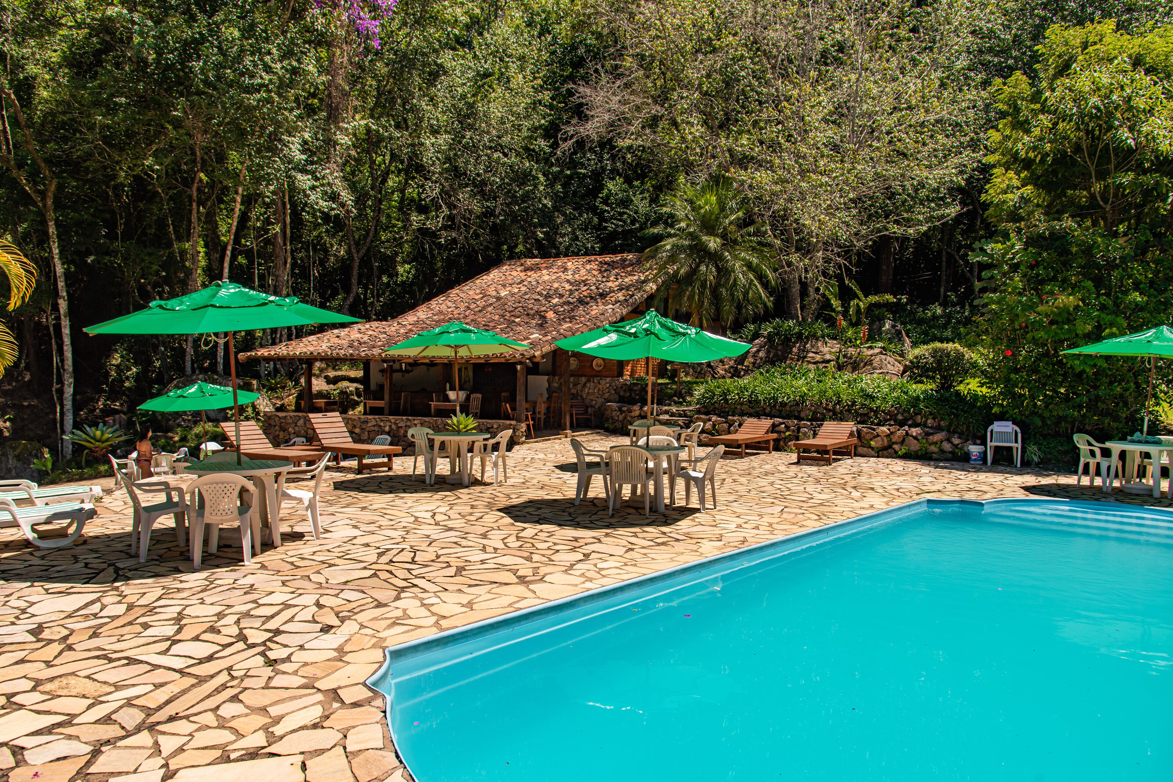 Uma área de piscina com piso de pedra, cadeiras de sol e guarda-sóis verdes.