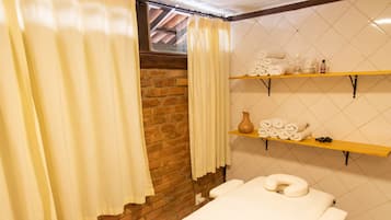 Um quarto de spa com mesa de massagem, parede de tijolos, prateleira de madeira com toalhas e janela com cortinas.