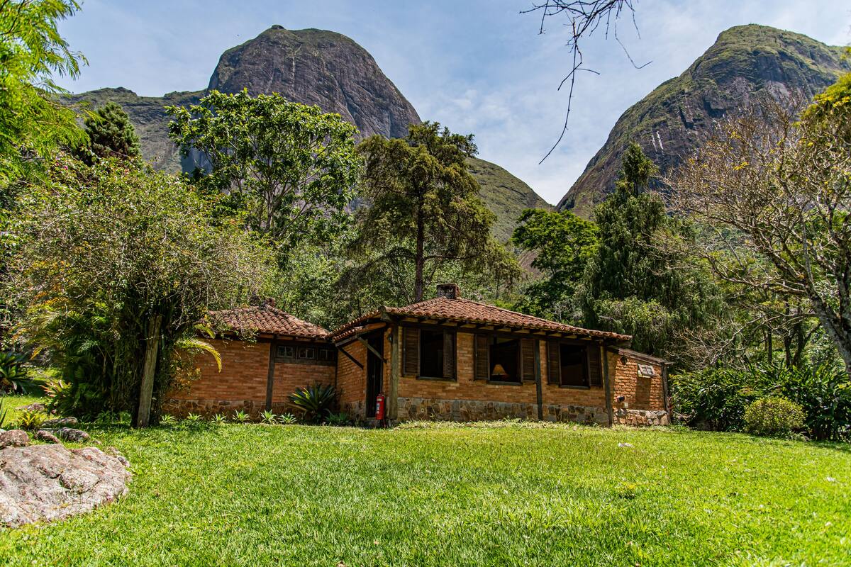 Una casa de ladrillo rústica con tejado de baldosas, rodeada de abundante vegetación y montañas al fondo.