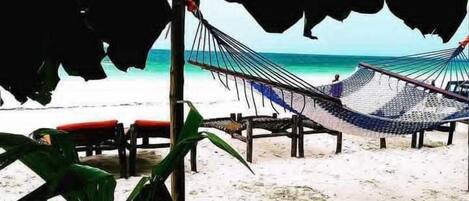 Uma praia com uma rede e cadeiras em área sombreada.