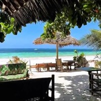 Un área de estar frente a la playa, con una sombrilla de paja, muebles de madera y vista al océano.