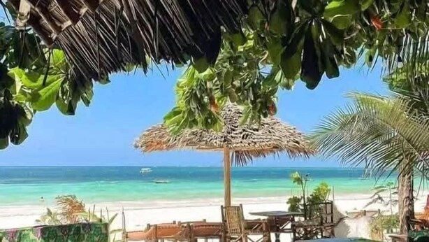 Ein Sitzbereich am Strand mit reetgedeckten Sonnenschirmen, Holzmöbeln und Blick auf das Meer.