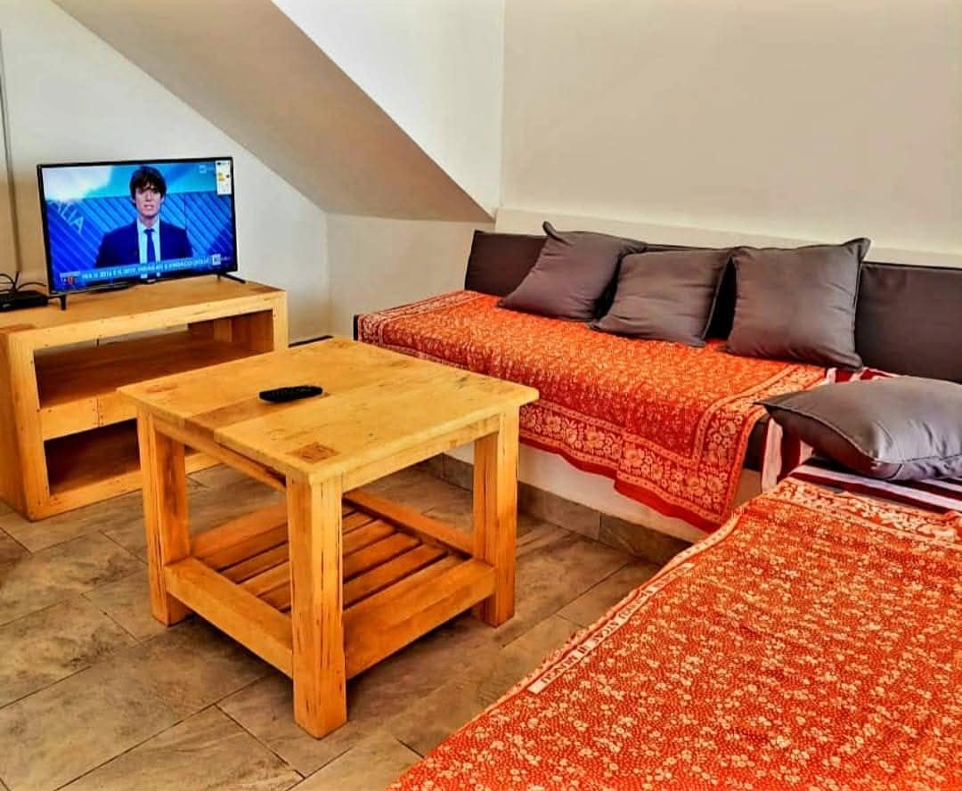Een kamer met een televisie op een houten standaard, een houten tafel, een bank met gebloemde dekens en een vloerkleed met dessin.