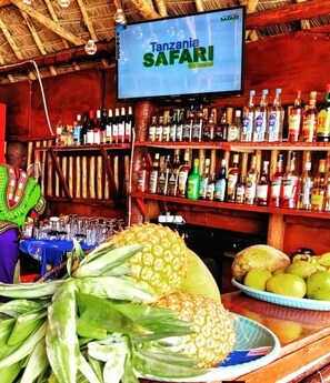Un bar doté d’un écran de télévision diffusant une émission sur les « safaris en Tanzanie » et de plusieurs étagères remplies de bouteilles d’alcool de toutes sortes.