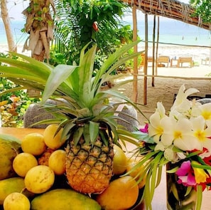 Un ambiente tropicale con vista sulla spiaggia, una struttura con tetto di paglia e una varietà di frutta e fiori.