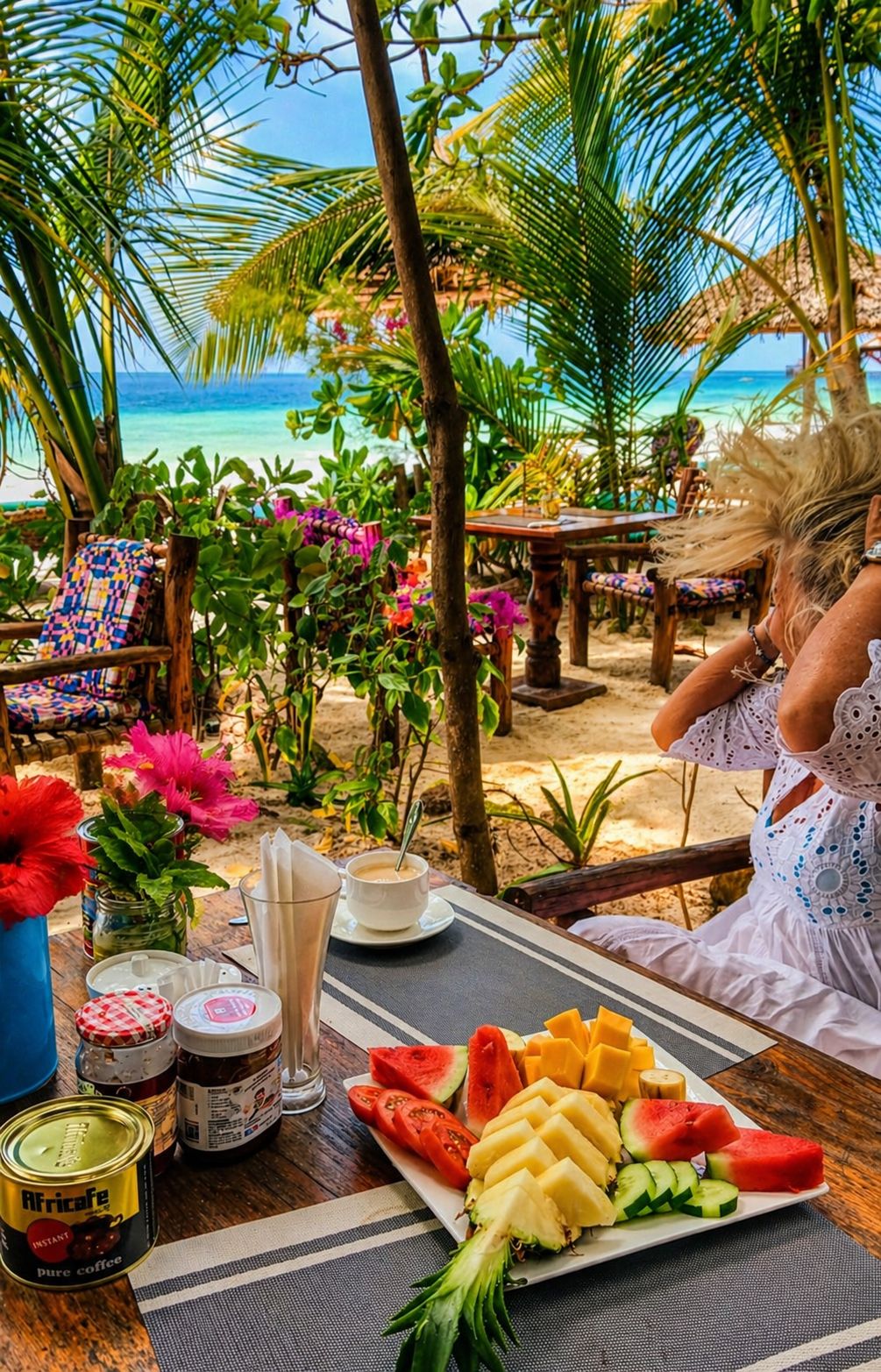 A tropical outdoor dining area with a view of the ocean, a table with food and drinks, and a person sitting at the table.