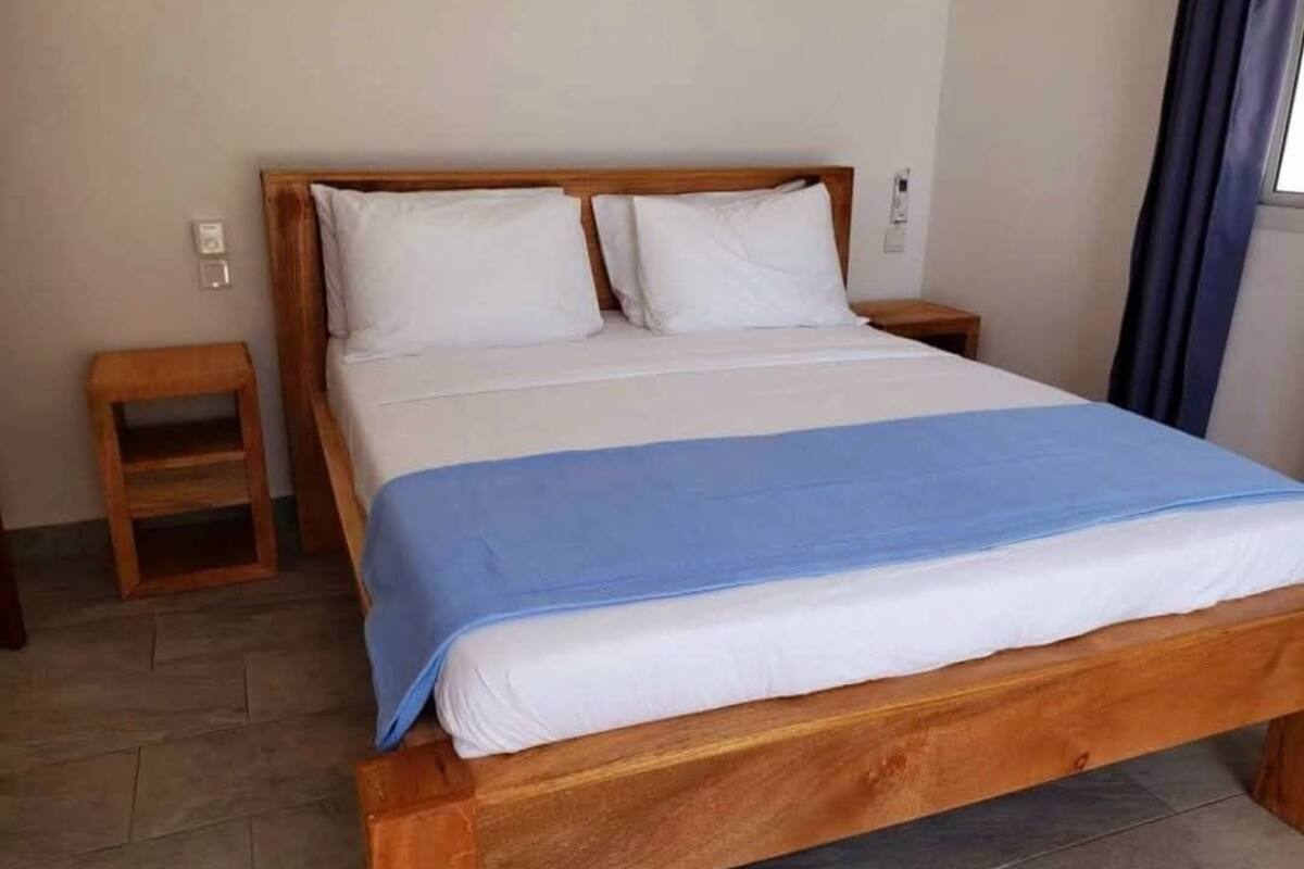 Habitació d'hotel estàndard amb un llit de fusta, dos coixins blancs, un edredó blau, una tauleta de nit i una taula de fusta petita.
