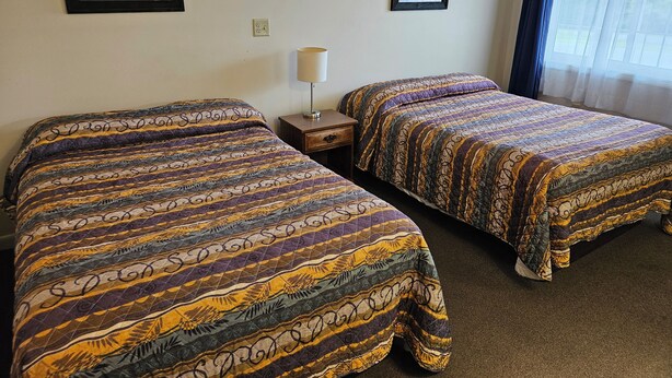 A hotel room with two beds, a nightstand, a lamp, and two framed pictures on the wall. - Brookside Inn (Smyrna Mills)