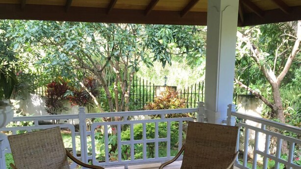 A covered patio with wicker furniture and a small table, surrounded by a white railing and lush greenery. - Ka Mario Guesthouse (Kandy)