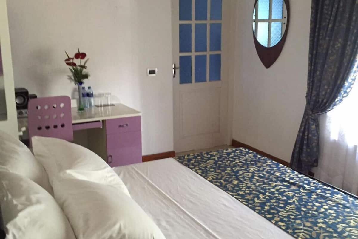A bedroom with a bed, a desk with a chair, a door with a window, and a curtain.