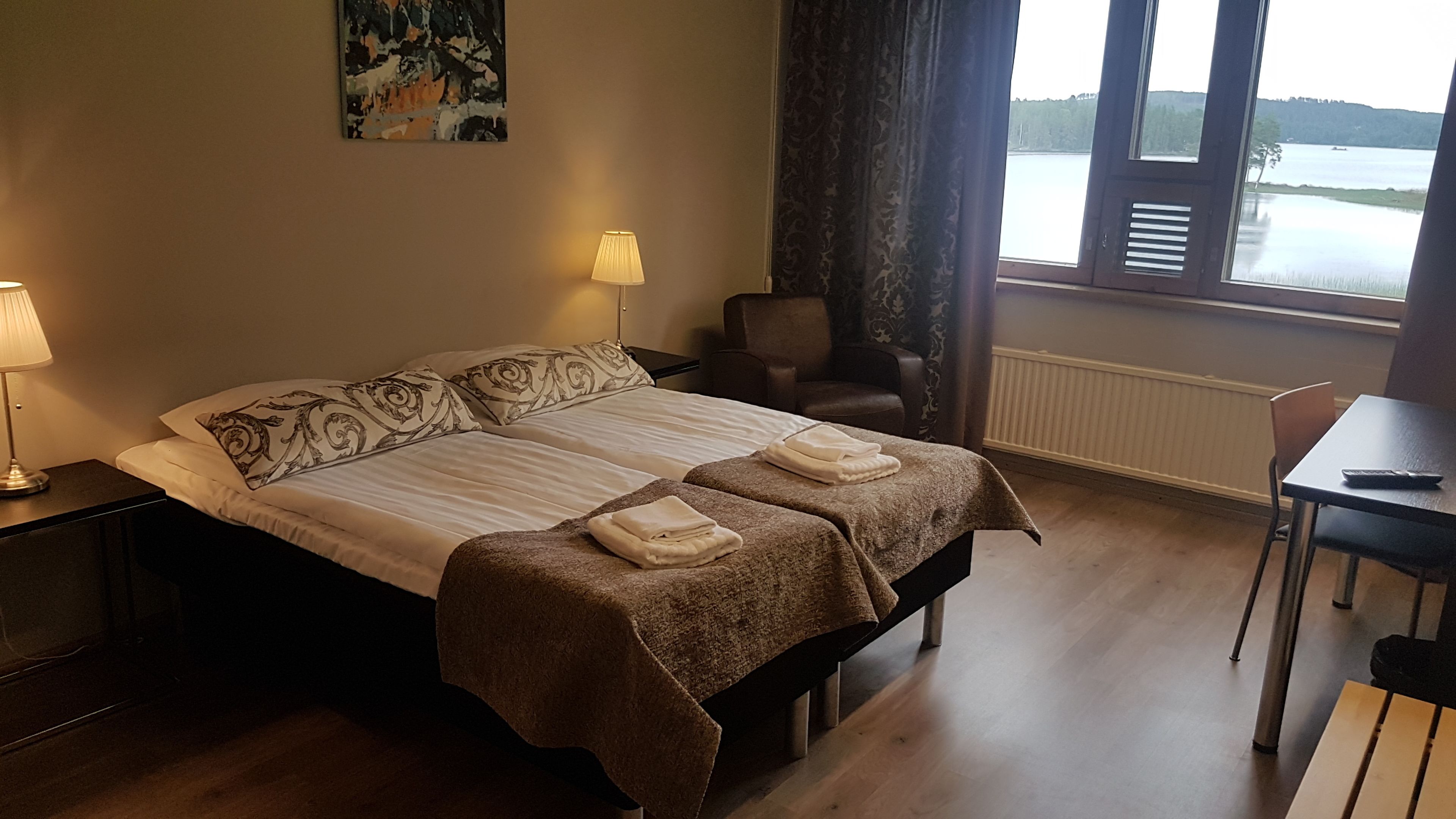 Hotellihuone, jossa on sänky, kaksi yöpöytälamppua, tuoli, pöytä ja ikkuna, josta avautuu näkymä järvelle.