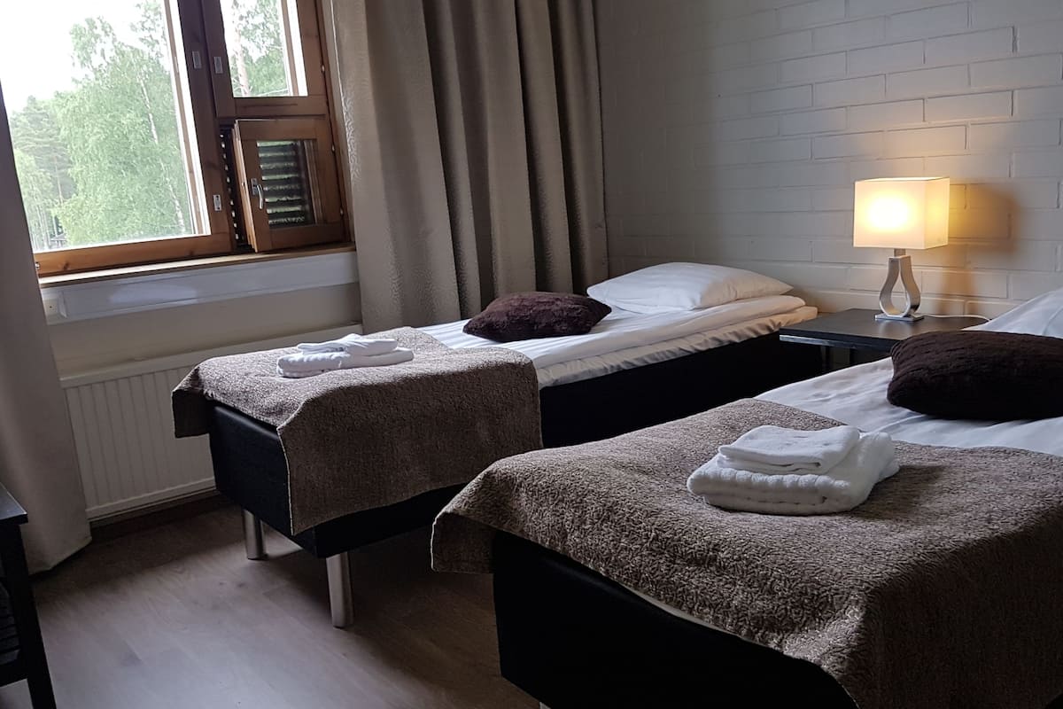 A room with two beds, each with a brown blanket and pillows, a bedside table with a lamp, and a window with curtains.