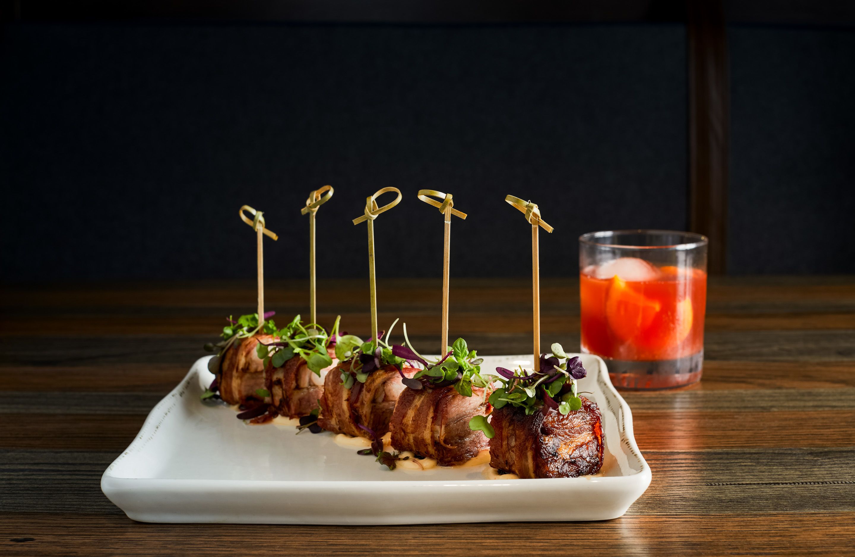 A white rectangular plate with skewered meat garnished with greens, accompanied by a glass of red beverage with ice cubes.