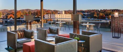 A modern outdoor seating area with leather sofas and tables, large windows offering a view of autumn trees and buildings.