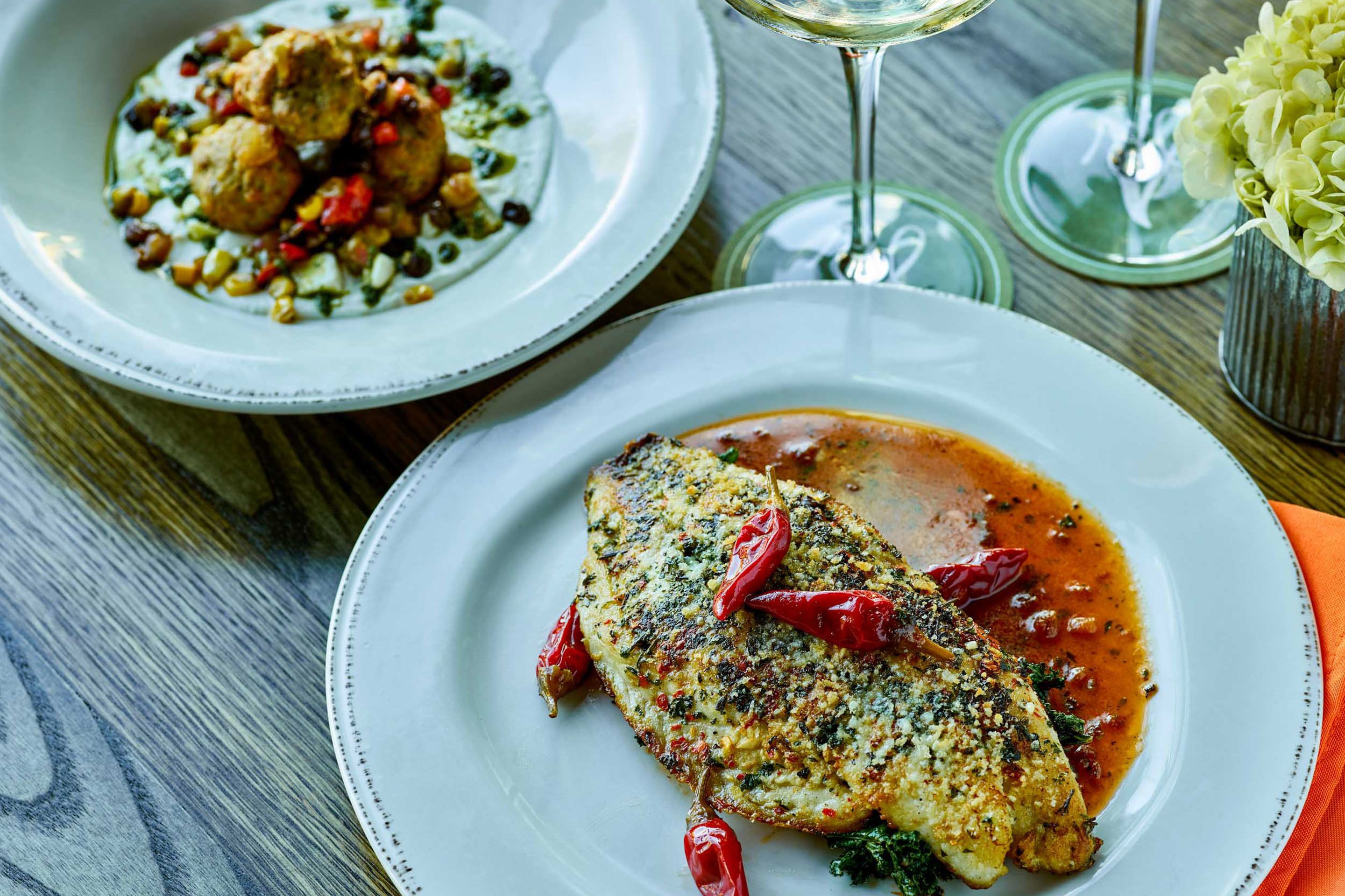 A dining table with two plates of food, one with a fish dish and the other with a meat dish, and a glass of white wine.