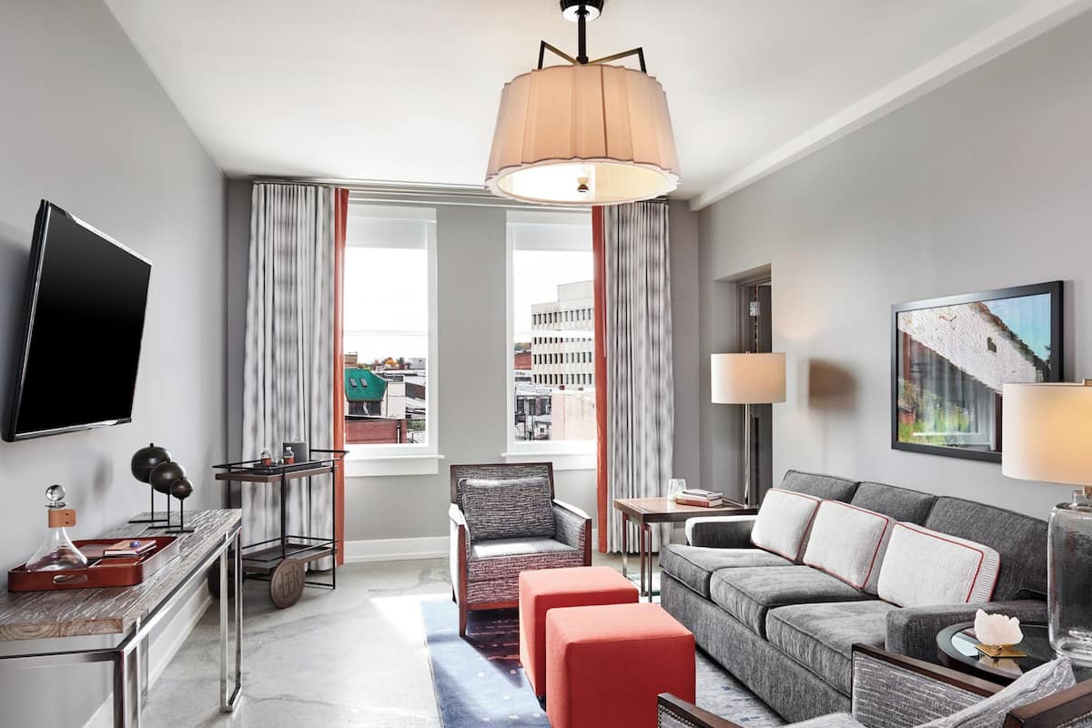 Una habitación de hotel moderna con un televisor de pantalla plana grande, un sofá cómodo, una mesita y una silla. La habitación tiene ventanas amplias con cortinas, una lámpara de techo y vistas a la ciudad.