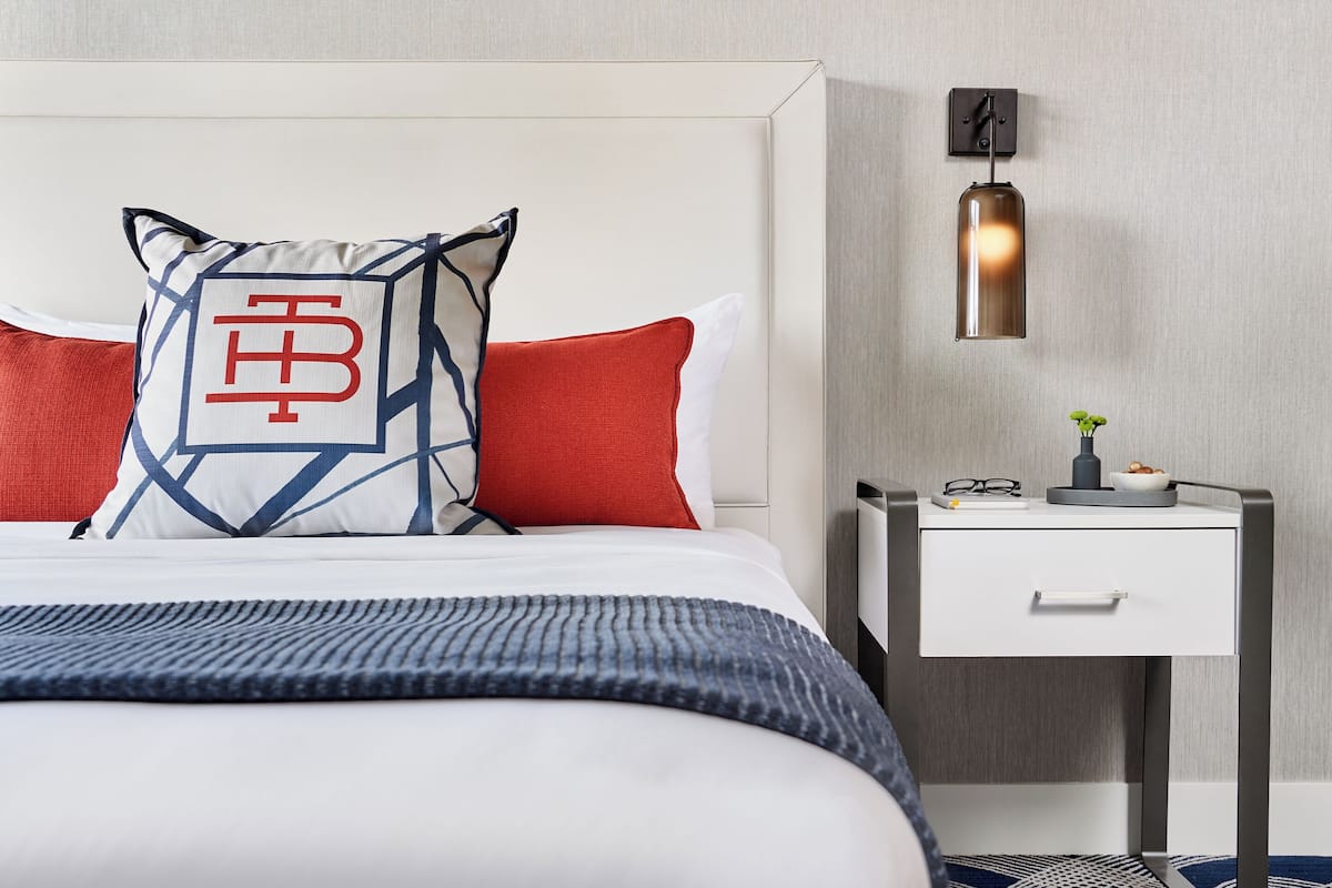Una cama bien hecha con una almohada estampada, una almohada roja y una manta azul. Hay una mesita de noche con una lámpara y una planta pequeña.