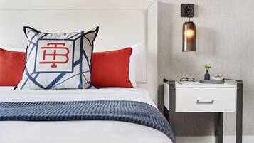 Een netjes opgemaakt bed met een gekleurd kussen, een rood kussen en een blauwe deken. Er staat een nachtkastje met een lamp en een klein plantje.