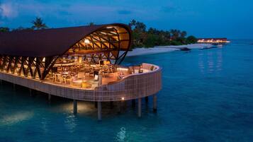 An overwater bungalows with a covered dining area, lounge chairs, and a view of the beach and ocean.