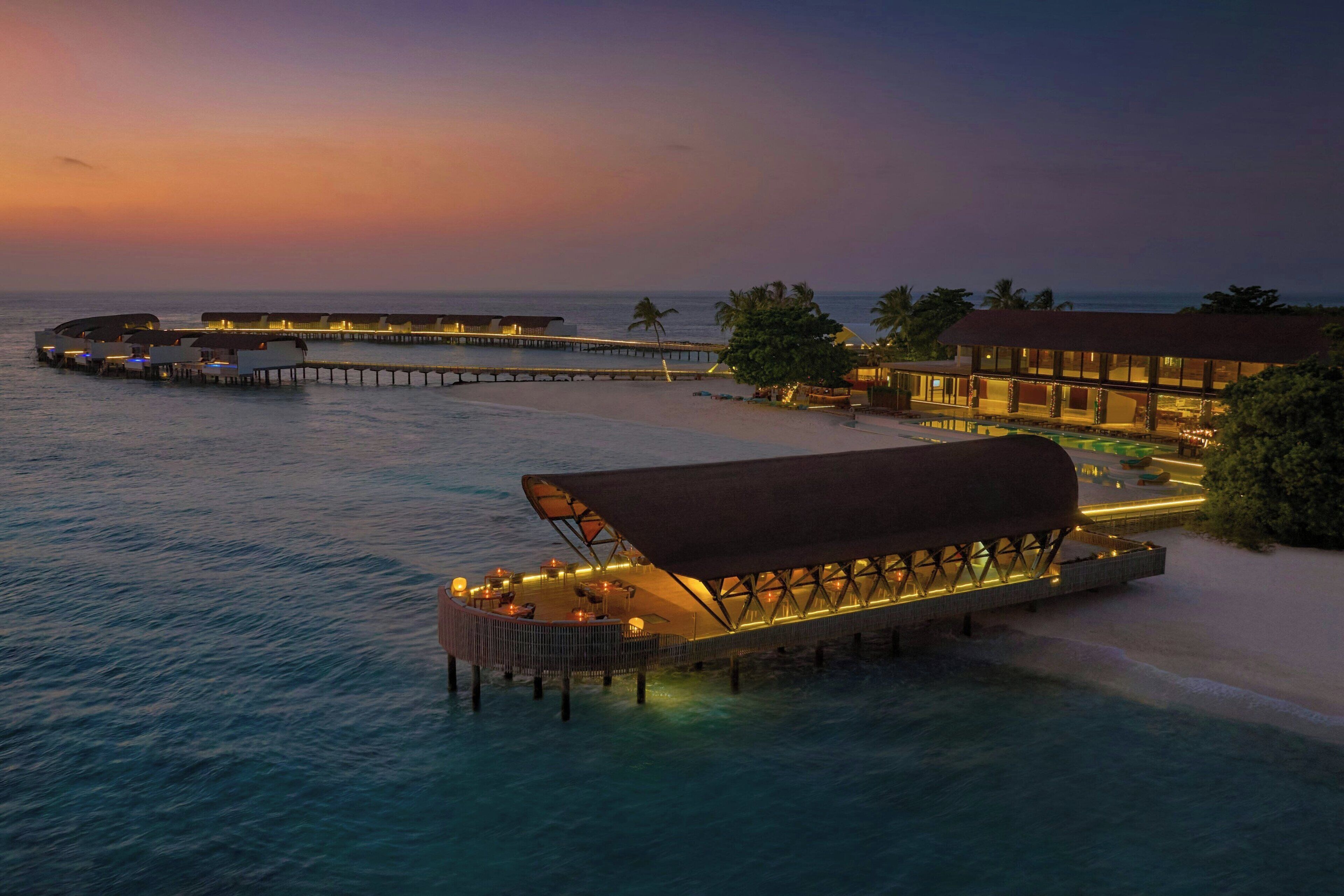 An overwater resort with a covered lounge area, a beach, and a pier extending into the ocean.