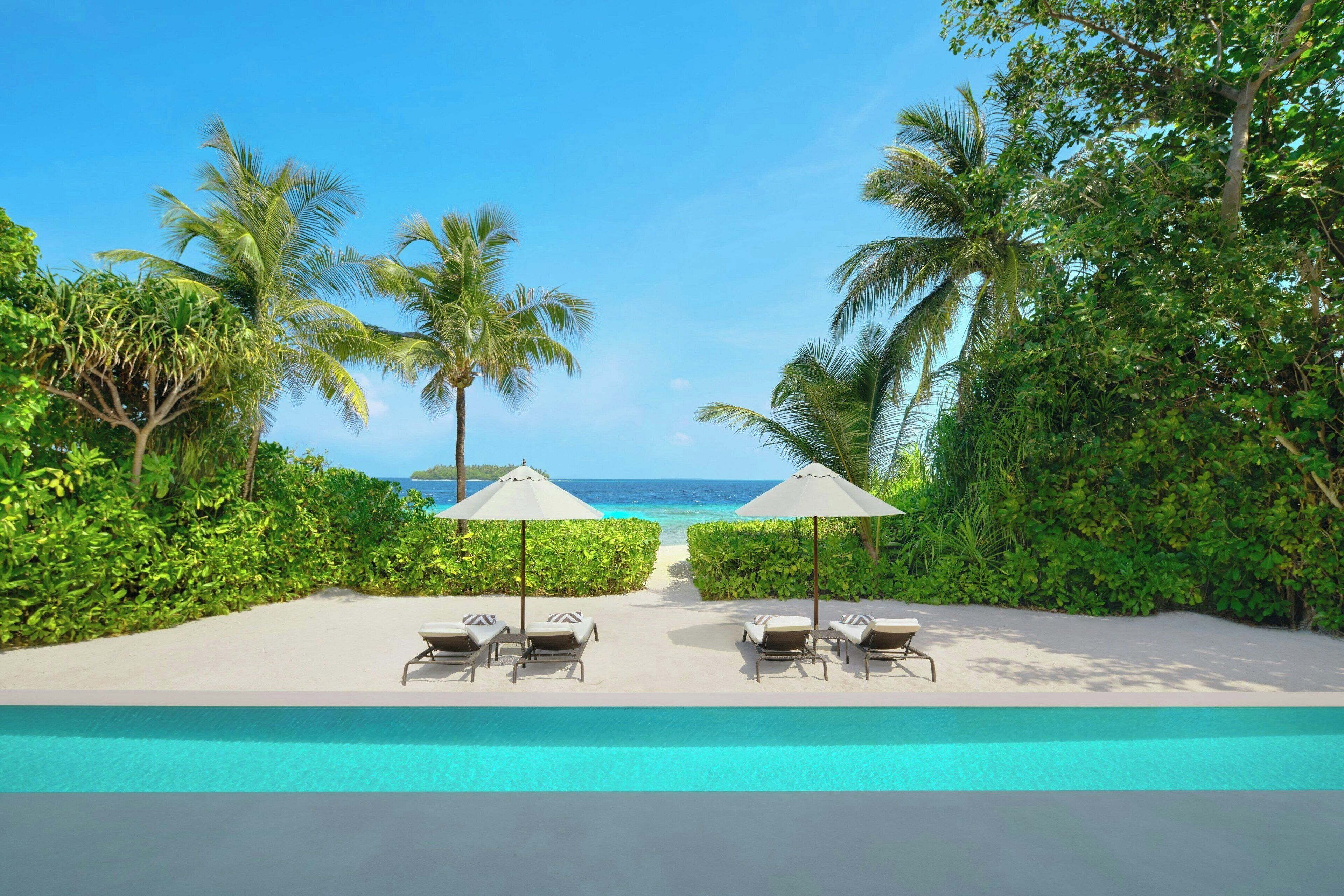 A poolside area with two lounge chairs and umbrellas, surrounded by lush greenery and a view of the ocean.