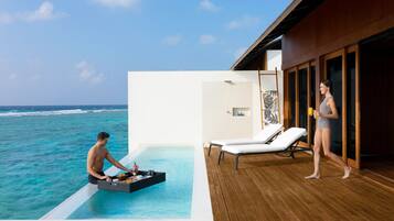 A man serving food from a tray into a pool, a woman walking by, and a lounge chair on a wooden deck with a view of the ocean.