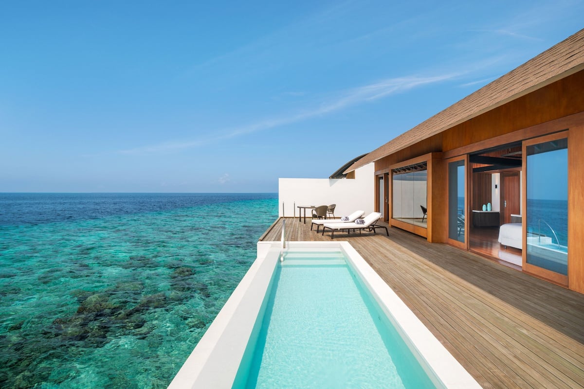 A modern infinity pool with a wooden deck, overlooking a clear blue ocean and coral reef.