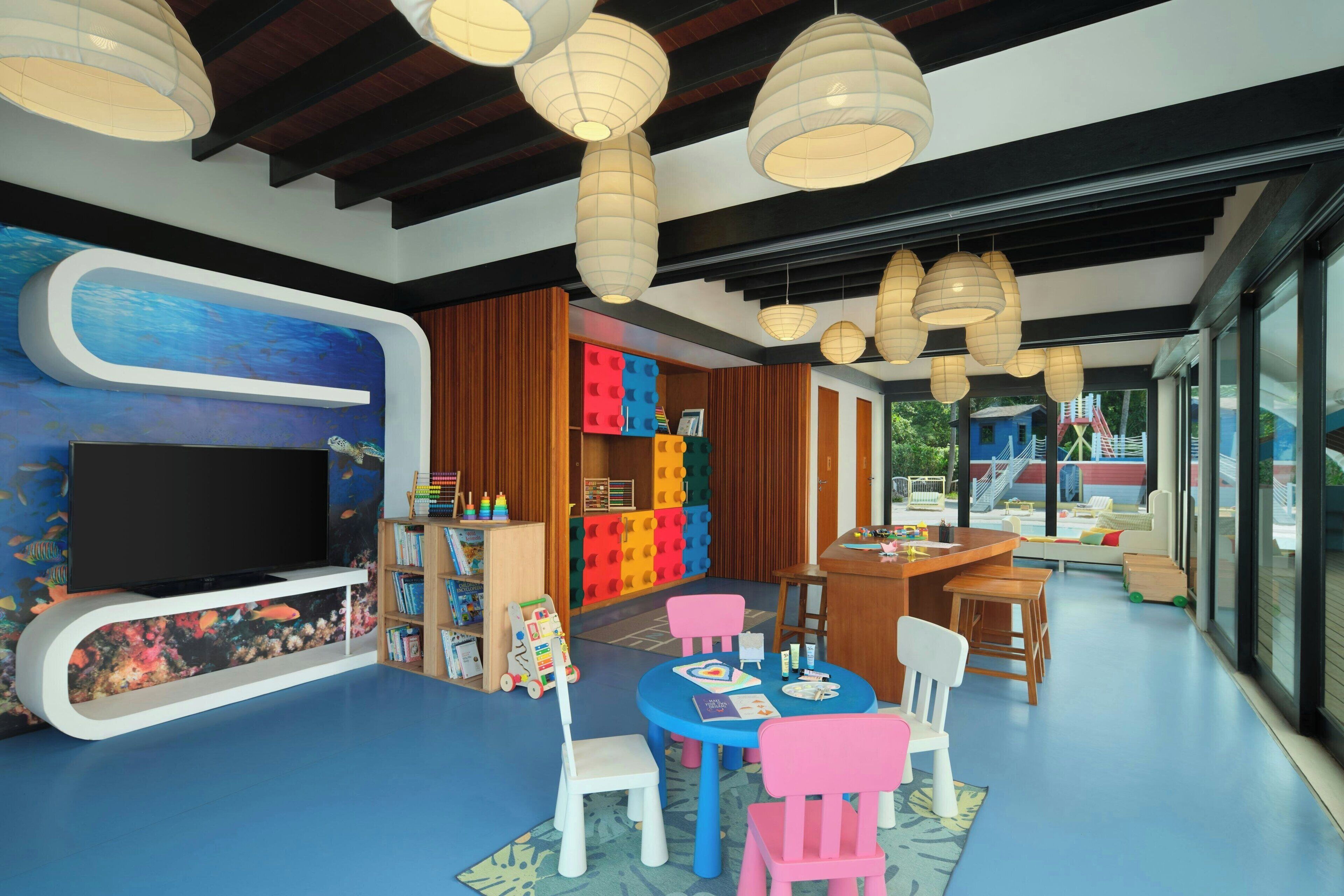 Un cuarto de juegos infantil con televisor, estructuras de juego coloridas y una zona de comedor con sillas rosas.