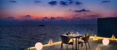 A sunset view from a wooden deck with a pool, dining table, chairs, and illuminated geometric light fixtures.