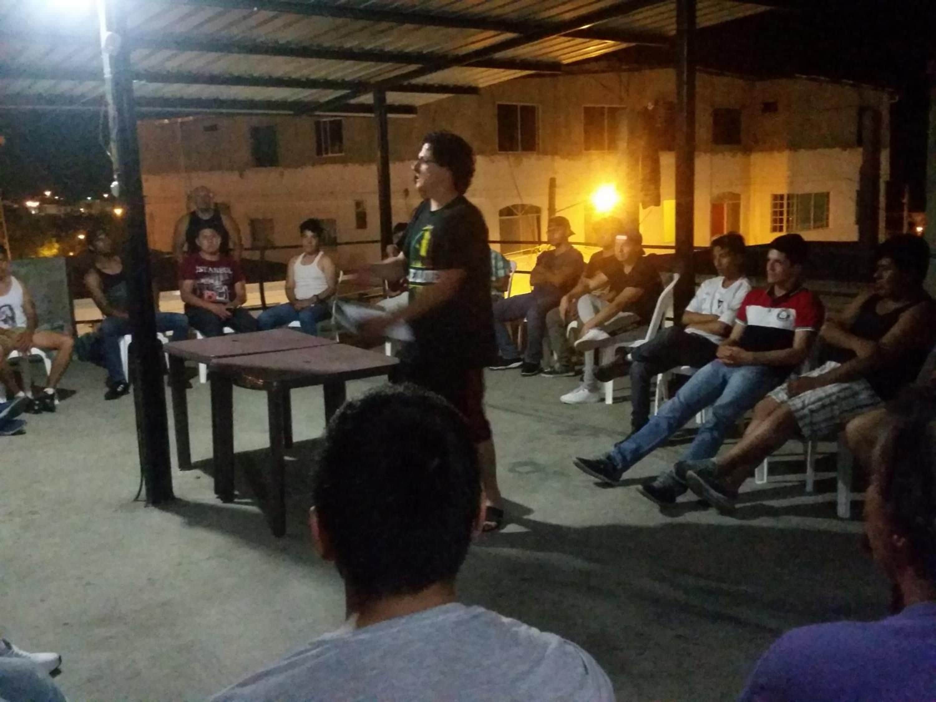 An outdoor gathering with people seated on plastic chairs under a metal roof structure.