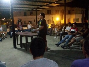 An outdoor gathering with people seated on plastic chairs under a metal roof structure. - Hostal El Naufrago (Manta)