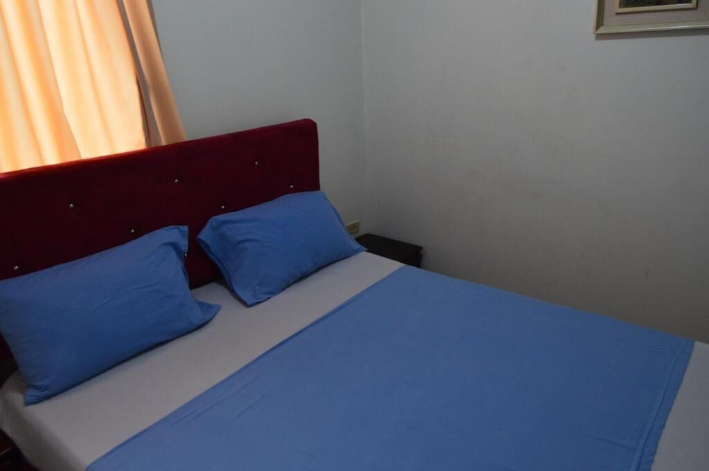 A single bed with a red headboard and blue bedding in a room with white walls and a window with curtains.
