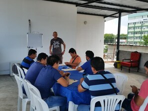 A group of people seated around a table in an outdoor setting with a whiteboard, chairs, and a building in the background. - Hostal El Naufrago (Manta)