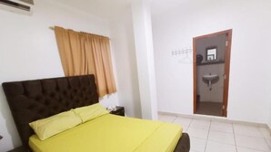 Un dormitorio con cama, mesita de noche, ventana con cortinas y baño visible a través de una puerta abierta.
