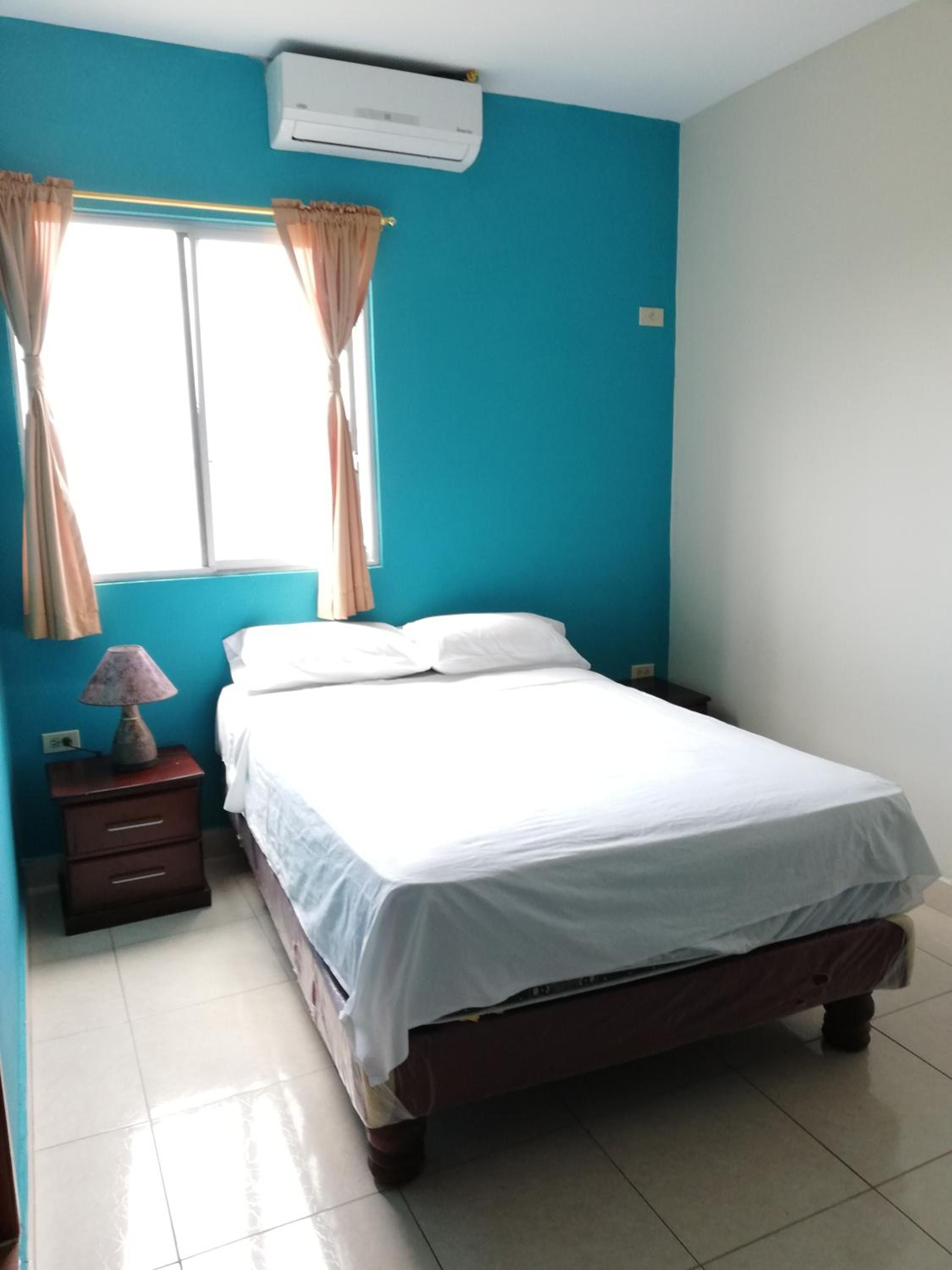 Un dormitorio con una cama, una mesita de noche, una lámpara, una ventana con cortinas y un equipo de aire acondicionado.