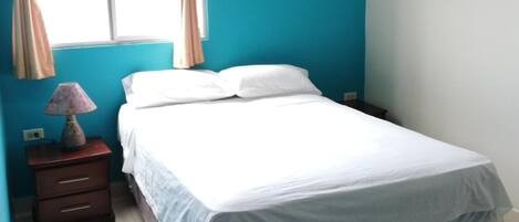 Un dormitorio con una cama, una mesita de noche, una lámpara, una ventana con cortinas y un equipo de aire acondicionado.