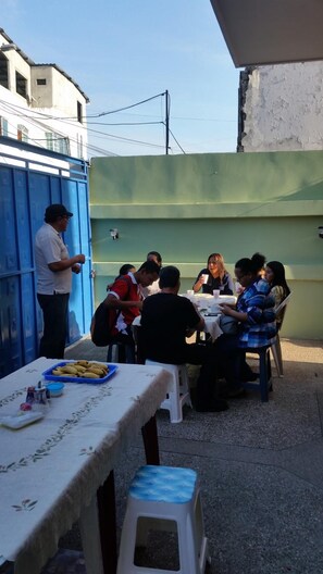 Reunião ao ar livre com pessoas sentadas em mesas, um recipiente azul com comida e uma toalha de mesa branca.