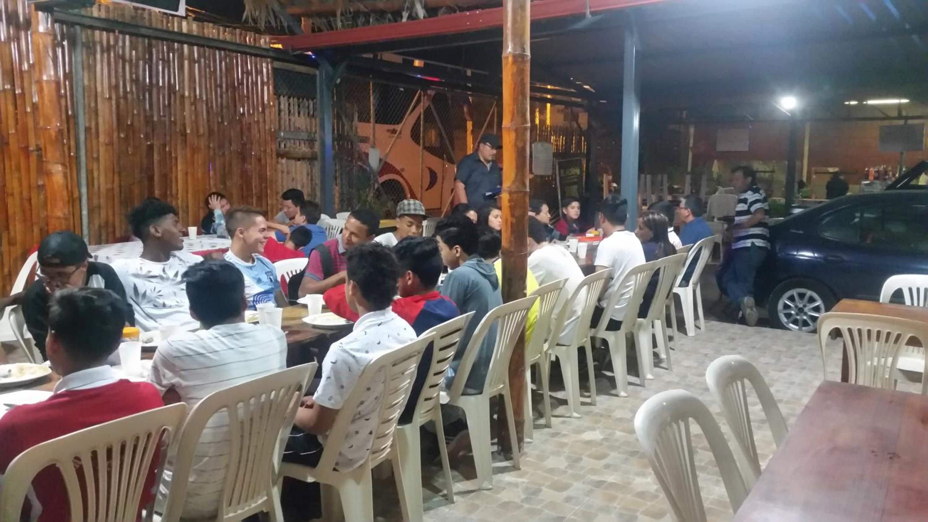 A crowded outdoor dining area with plastic chairs and tables, surrounded by a bamboo fence.