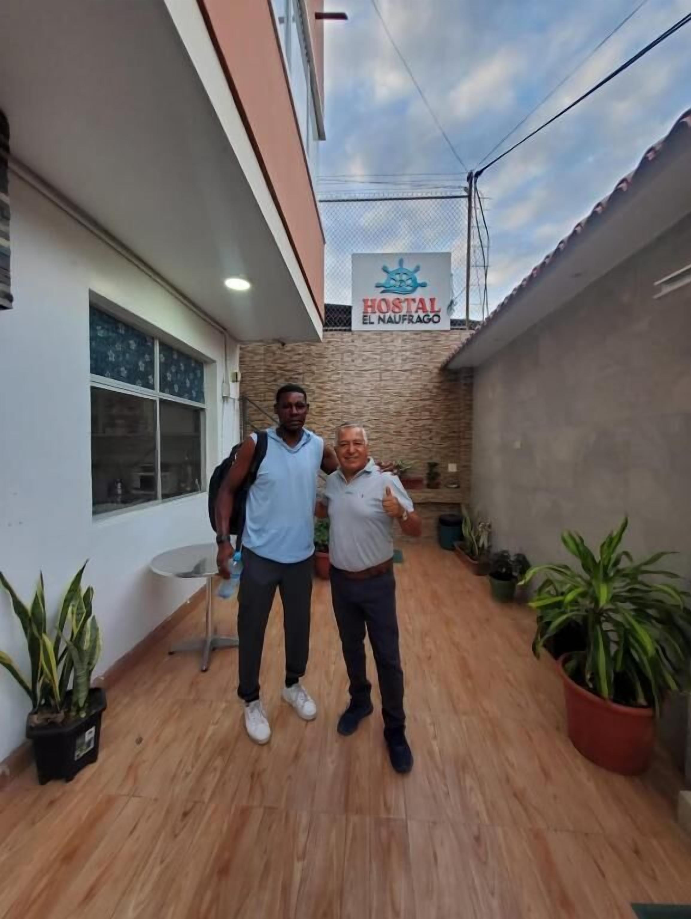Two individuals standing in a courtyard with a sign for 'Hostal El Naufrago' above.