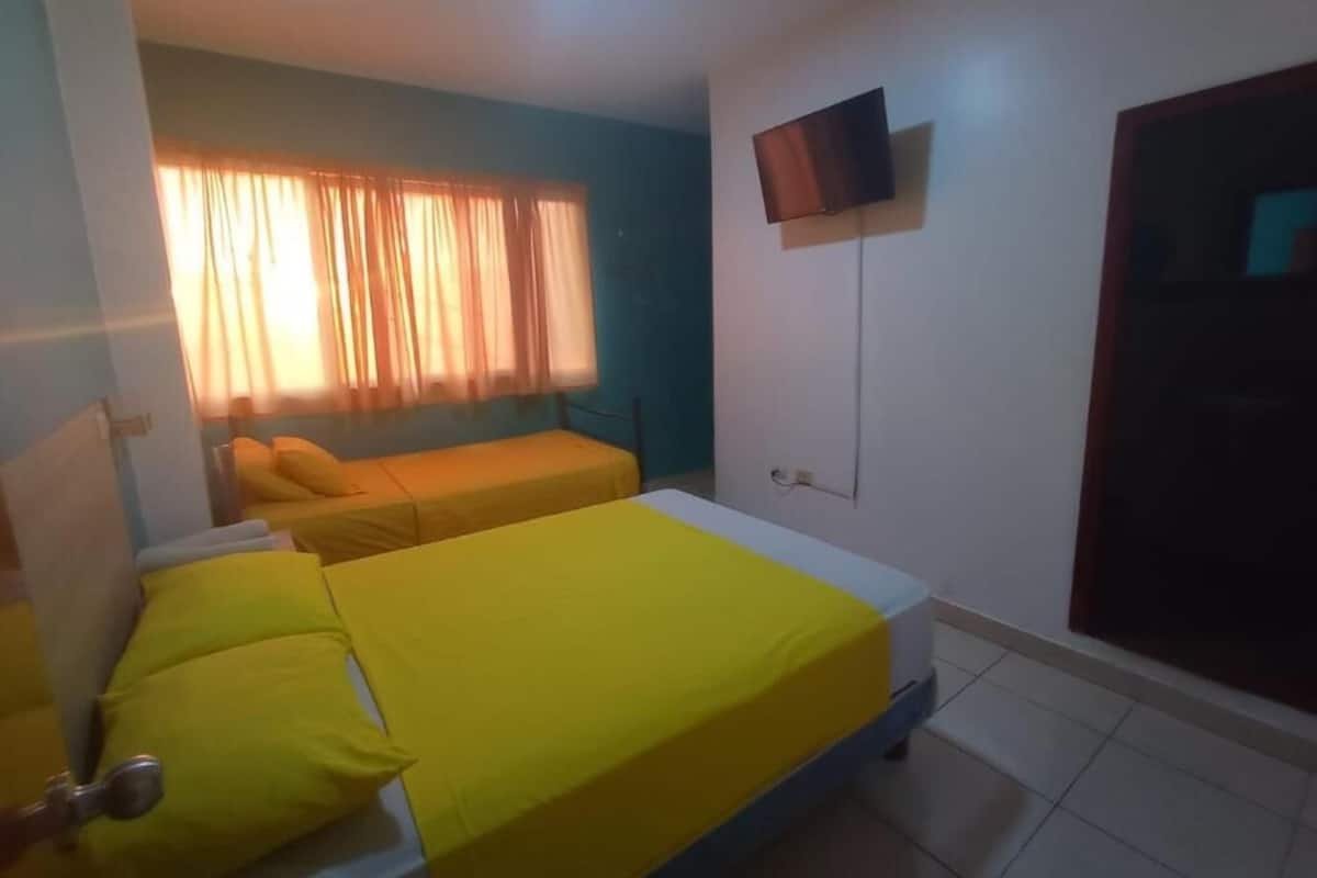 A room with two beds, a television mounted on the wall, and a window with curtains.