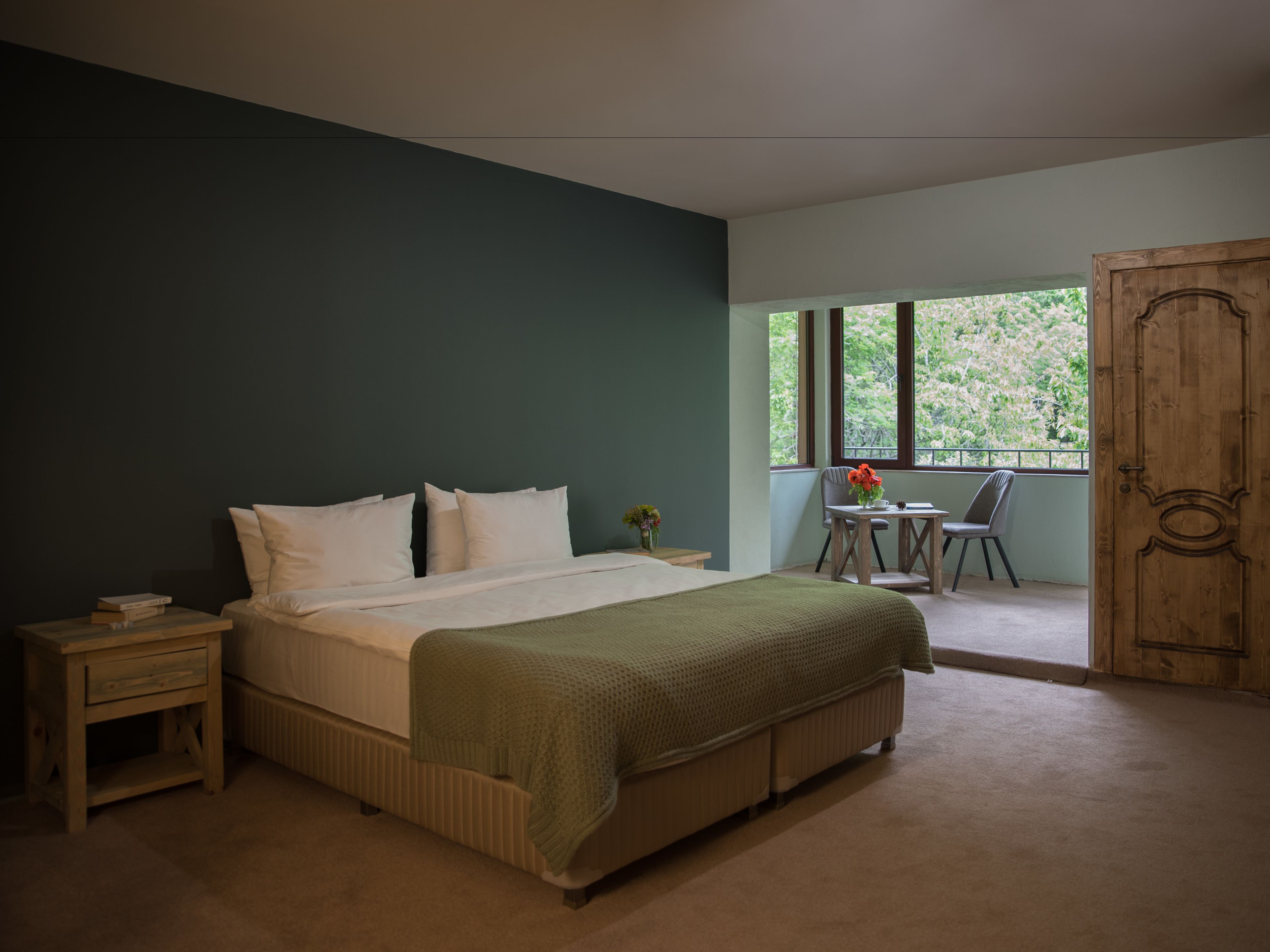 Une chambre à coucher avec un lit, des tables de chevet, une fenêtre donnant sur un paysage verdoyant et une porte en bois.