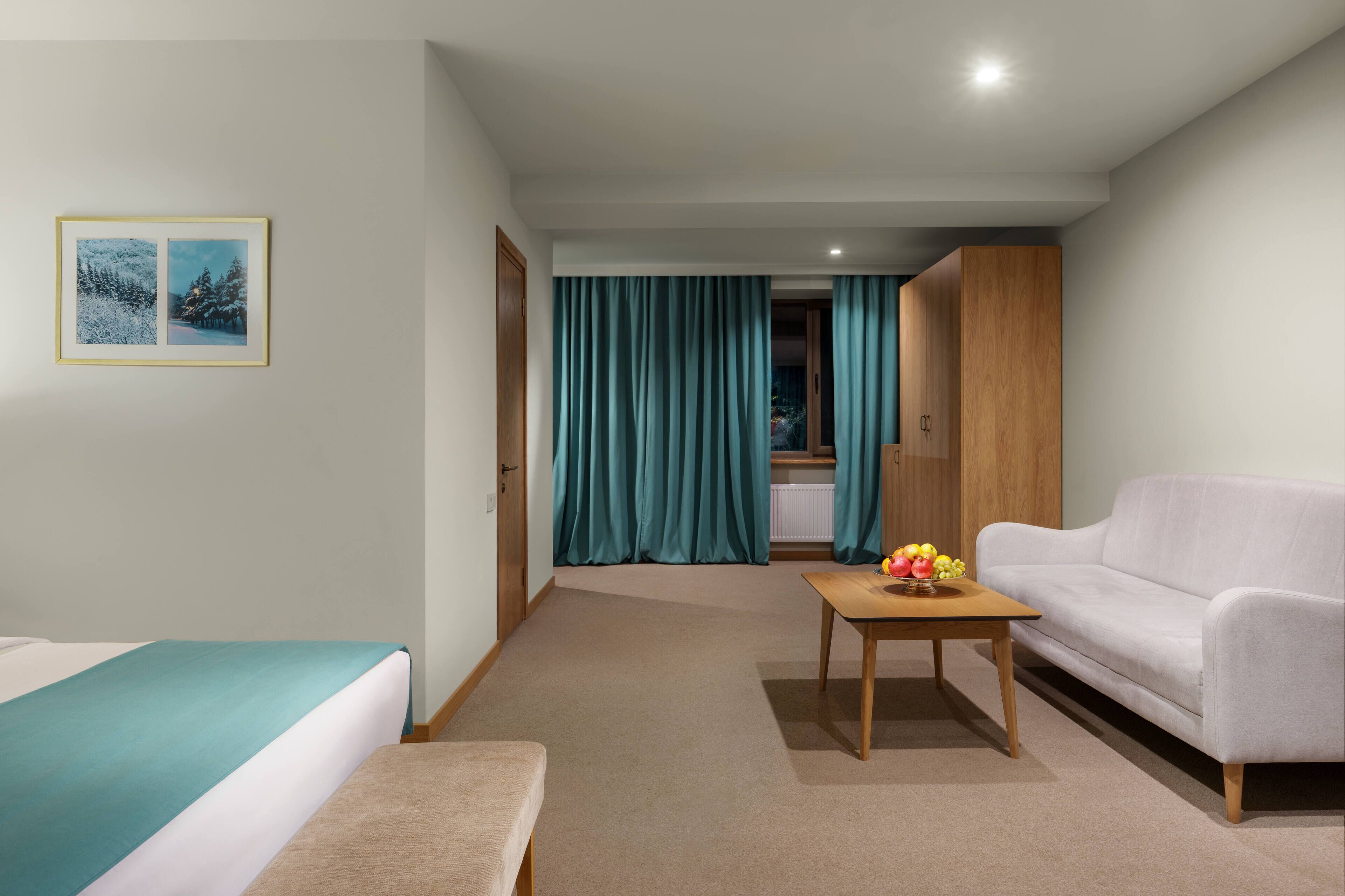 A hotel room with a bed, a sofa, a small table with fruit, teal curtains, and a framed picture on the wall.