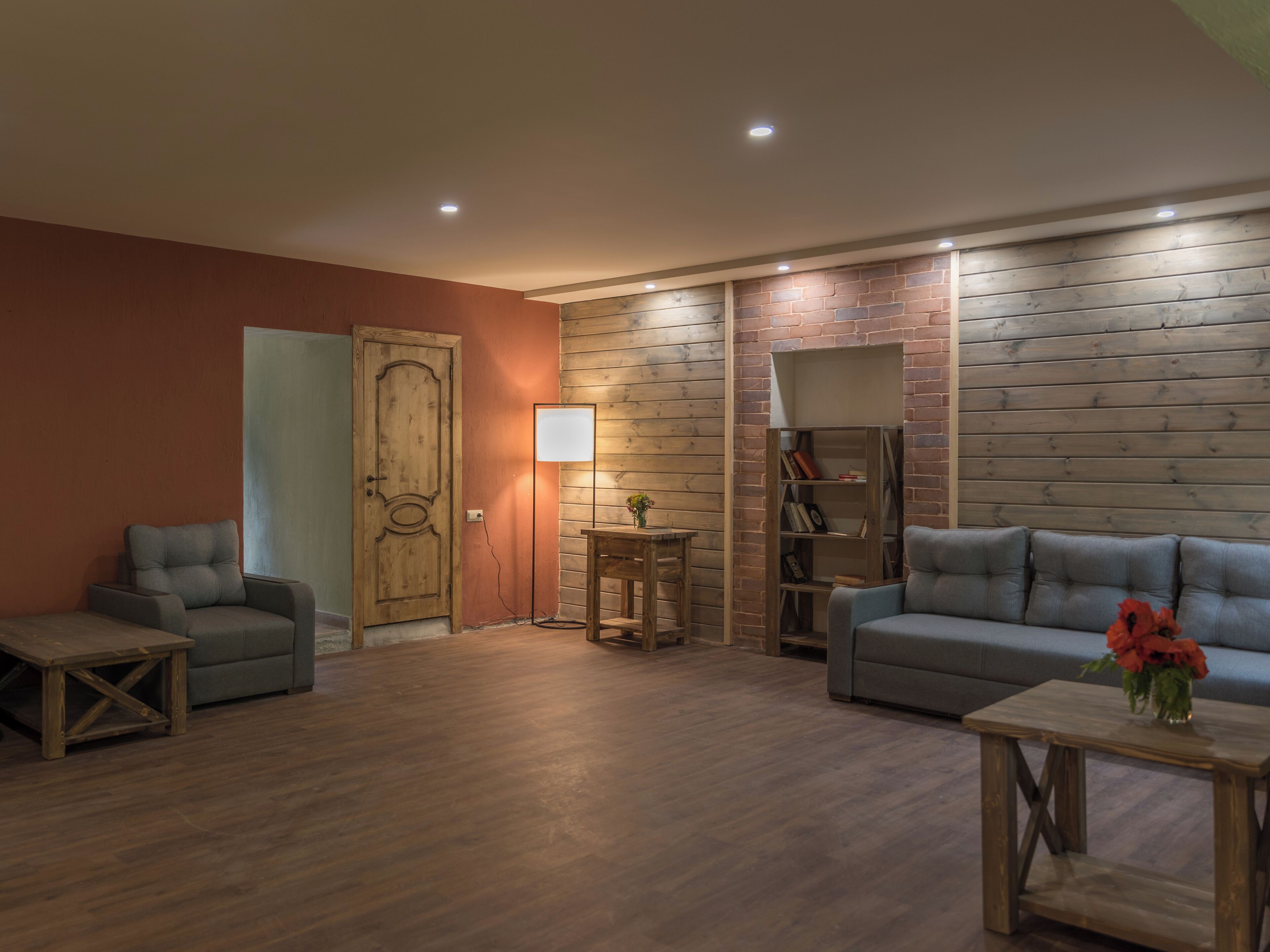 A cozy living room with a wooden floor, a grey sofa, a wooden coffee table, and a bookshelf against a brick wall.