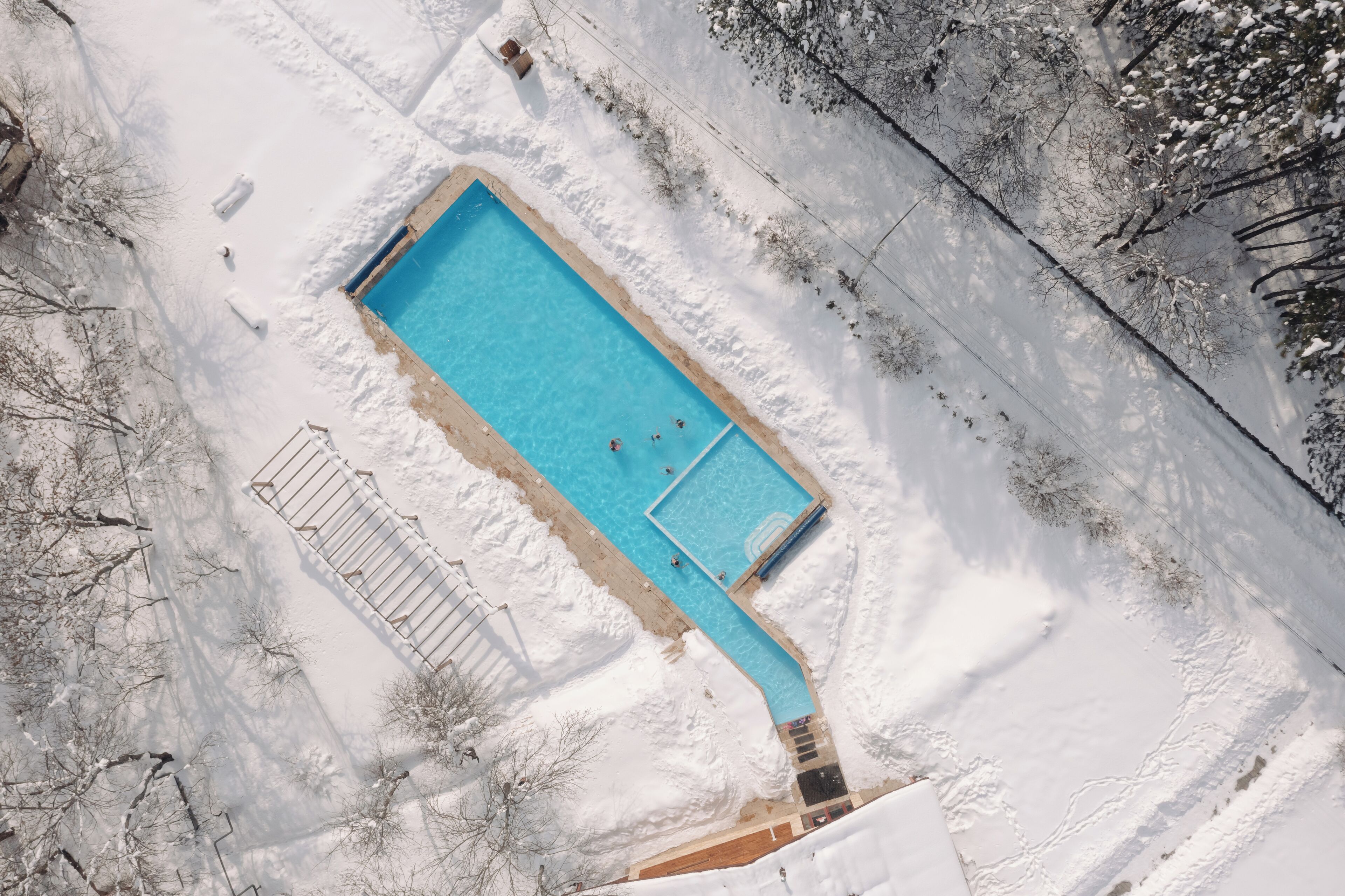 Vista aérea de una piscina rectangular rodeada de nieve, con algunas personas nadando.