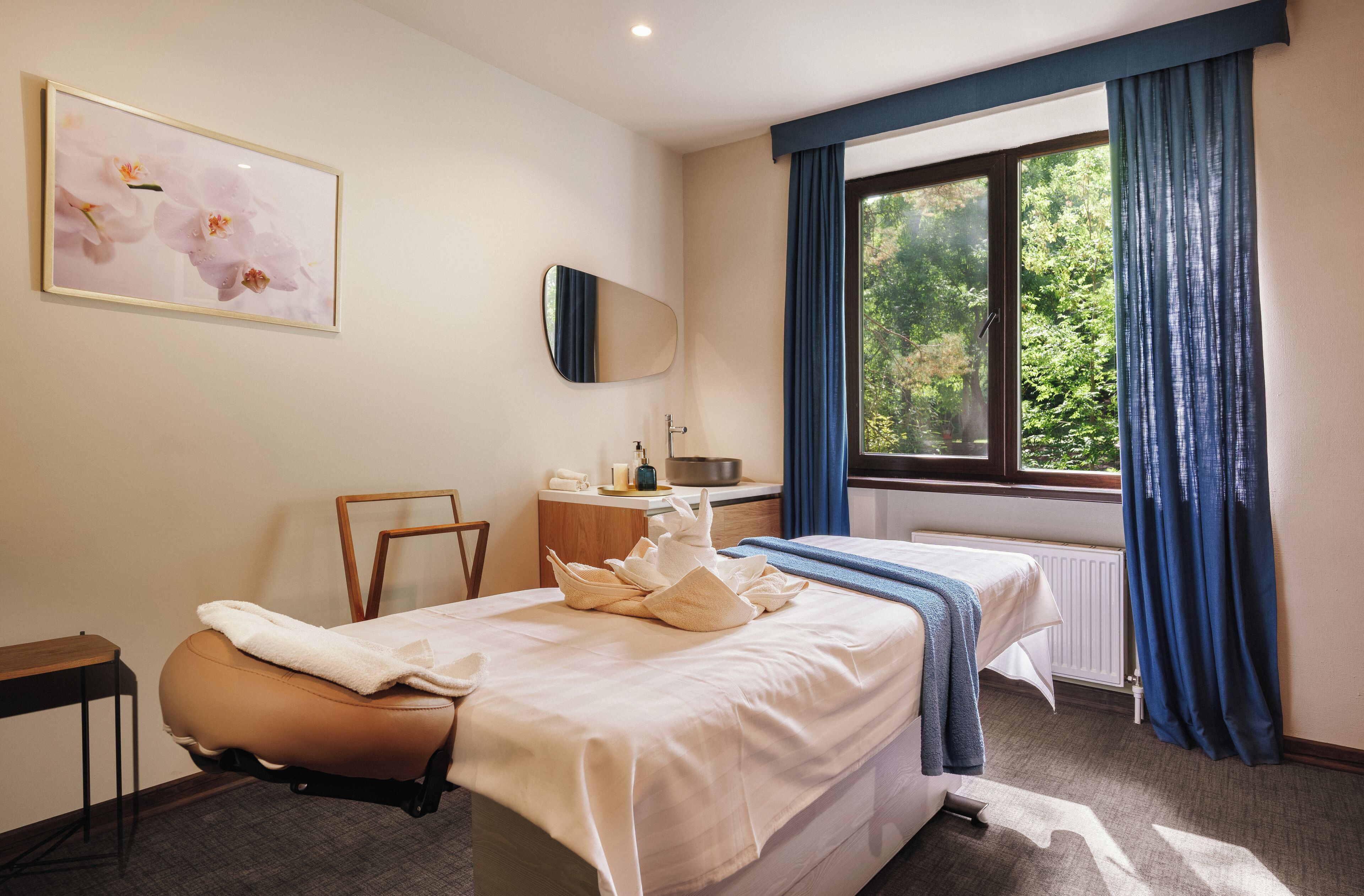 Habitación de hotel con cama, escritorio, silla, ventana con cortinas, radiador y un cuadro en la pared.