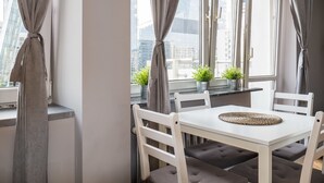 A modern dining area with a white table and chairs, large windows with curtains, and potted plants on the windowsill. - 55 Plater Residence (Warsaw)