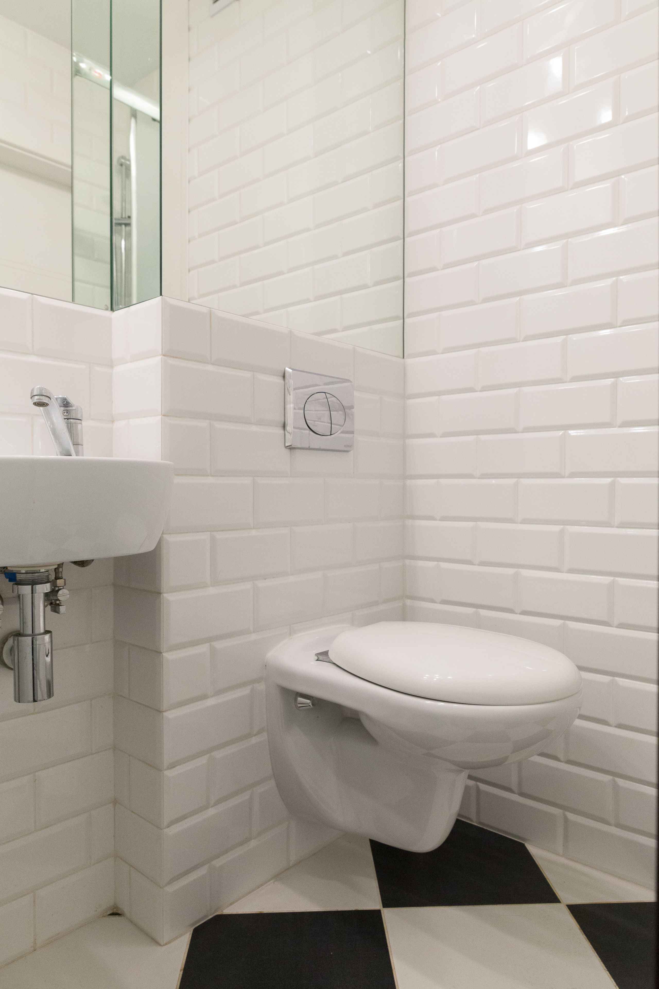 Un baño con azulejos blancos tipo metro, un inodoro blanco, un lavabo con grifo cromado y un suelo a cuadros blanco y negro.