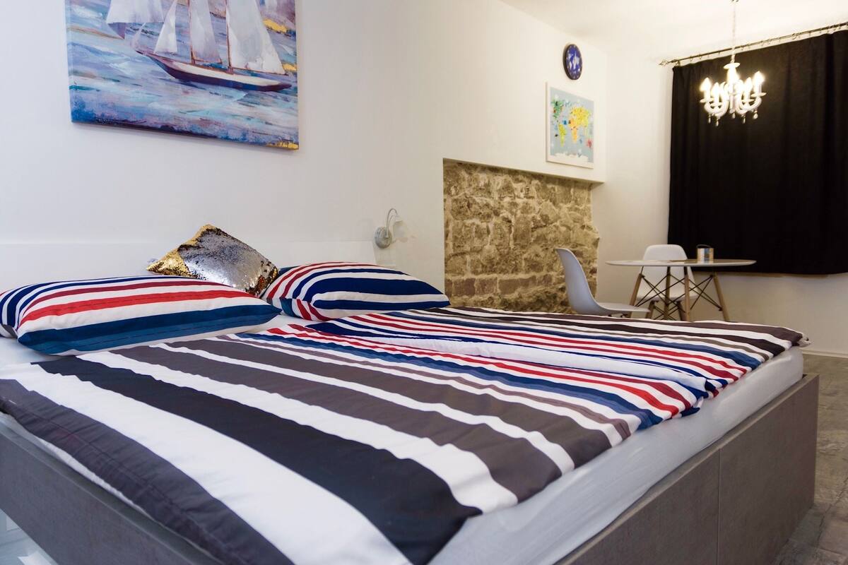 Una cama con ropa de cama a rayas, una pared de piedra, una pequeña mesa con sillas y un candelabro.