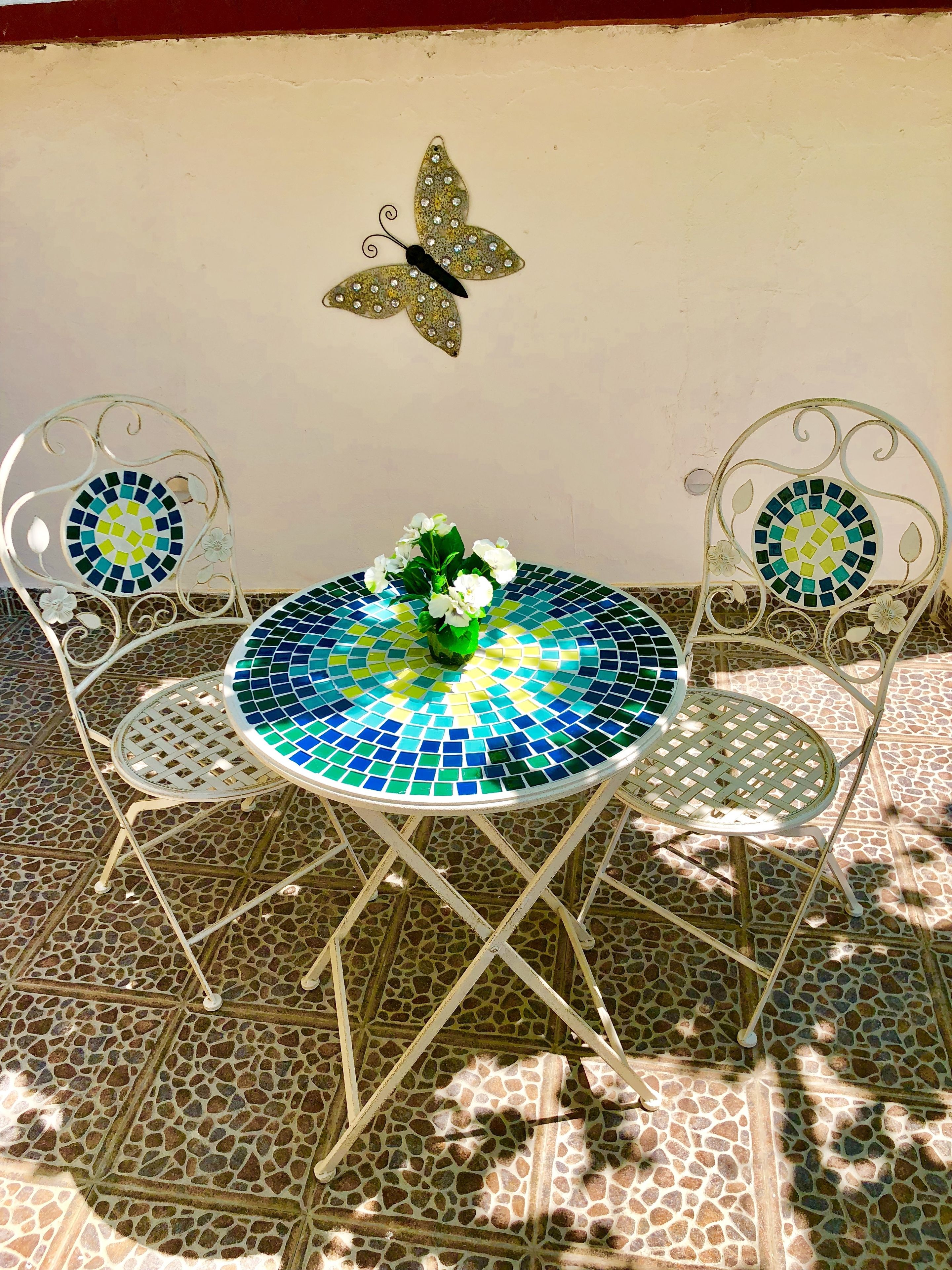 Una mesa redonda con cubierta de mosaico, dos sillas de diseño similar y una decoración de mariposa en la pared.