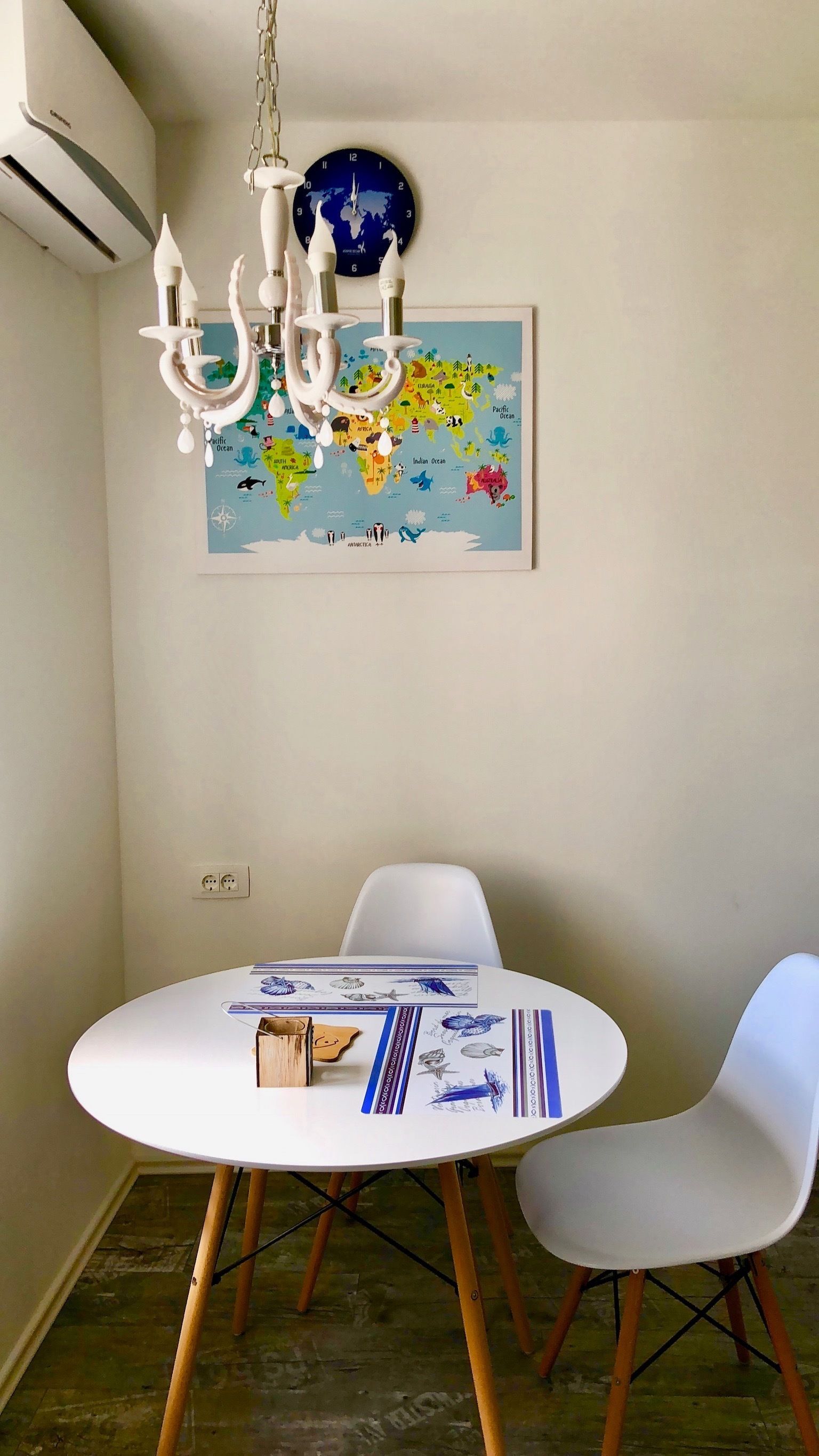 A dining area with a round table, two white chairs, a chandelier, a world map on the wall, and an air conditioning unit.